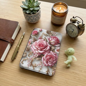 May include: A decorative phone case featuring a floral design with pink roses and gold accents. The image also includes a lit candle, a small alarm clock, a succulent plant, a pen, a notebook, and a small figurine.