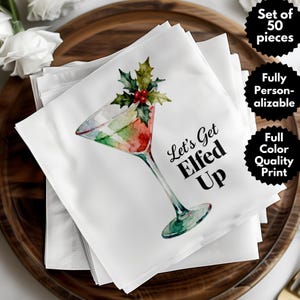 May include: White cocktail napkins with a watercolor illustration of a martini glass decorated with holly and berries. The text "Let's Get Elfed Up" is printed on the napkins. The napkins are stacked on a wooden tray.