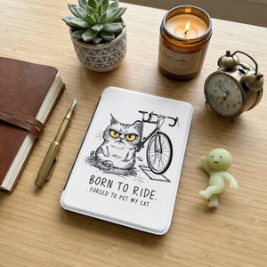 May include: A white tablet case with a grumpy cat illustration next to a bicycle. The text "BORN TO RIDE. FORCED TO PET MY CAT." is below the illustration. Other items include a candle, a succulent, a clock, a pen, a notebook, and a small figurine.