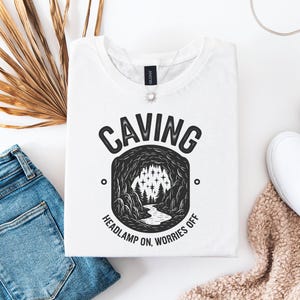 Puede incluir: Camiseta blanca con una ilustración en blanco y negro de una cueva con el texto "Caving Headlamp On, Worries Off" impreso en la parte delantera.
