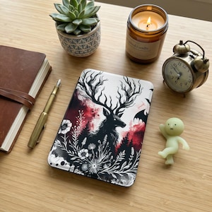 May include: A decorative phone case featuring a black and white illustration of a stag with large antlers, set against a backdrop of red and black. The case is surrounded by floral and forest elements. A small alarm clock and a succulent plant are also visible.