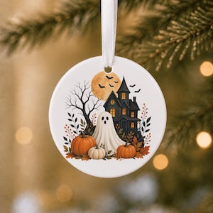 May include: A round white ceramic ornament hanging from a white ribbon. The design features a dark haunted house, a white ghost, orange pumpkins, autumn foliage, a large yellow moon, and black bats against a white background.