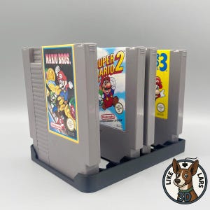 Könnte beinhalten: Drei graue Nintendo Entertainment System Spielkassetten stehen aufrecht in einem dunkelblauen Kunststoff-Aufbewahrungsständer. Die Kassetten zeigen Etiketten für Mario Bros., Super Mario Bros. 2 und Super Mario Bros. 3 mit bunten Illustrationen.
