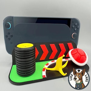 Op de afbeelding: Een zwart en grijs gameconsole-dock met een 3D-geprint ontwerp met racethema. Het ontwerp omvat een band, een bananenschil, een rode schildpadschild en een groene en zwarte baan. Het dock is ontworpen om een gameconsole te bevatten.