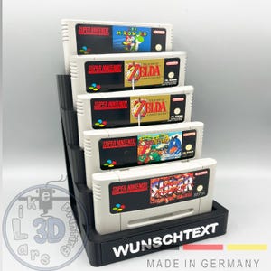 Könnte beinhalten: Schwarzer, mehrstufiger Display-Ständer mit fünf Super Nintendo-Spielkassetten. Die Kassetten zeigen Spieletitel wie "Super Mario World" und "The Legend of Zelda". Der Ständer trägt den Text "WUNSCHTEXT" und "MADE IN GERMANY".