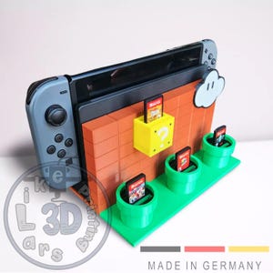 Puede incluir: Un soporte de impresión 3D para Nintendo Switch con un diseño de pared de ladrillo y soportes de cartuchos de juego en forma de tuberías verdes. El soporte presenta una nube y un bloque de signo de interrogación. "MADE IN GERMANY" está impreso en la base.
