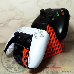 Peut inclure: Une manette PlayStation 5 blanche et noire et une manette Xbox noire reposent sur un support orange et noir en nid d'abeille. Le support est étiqueté "MADE IN GERMANY".