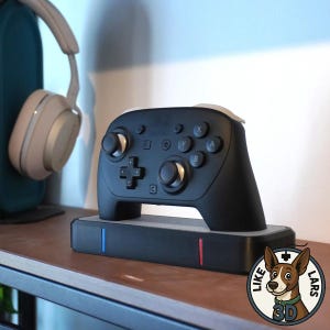 Può includere: Un controller di gioco nero con dettagli bianchi, appoggiato su una base di ricarica nera con strisce blu e rosse. Il controller ha i pulsanti X, Y, A e B. Sullo sfondo sono visibili delle cuffie beige.