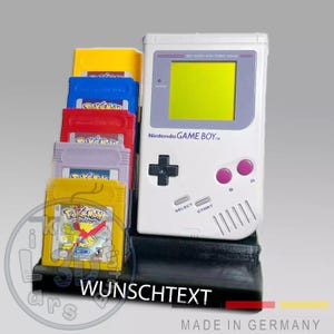 Könnte beinhalten: Ein weißer Nintendo Game Boy-Konsolen mit grünem Bildschirm und mehreren Spielkassetten in verschiedenen Farben. Die Kassetten sind in einem schwarzen Halter gestapelt. Der Text "WUNSCHTEXT" ist auf dem Halter sichtbar, und "MADE IN GERMANY" unten.