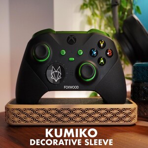 Könnte beinhalten: Ein schwarzer Gaming-Controller mit grünen Akzenten, einem weißen geometrischen Fuchsemblem und dem Schriftzug FOXWOOD. Der Controller steht auf einer hellen, rechteckigen Basis mit einem komplexen, sich wiederholenden Sternmuster.