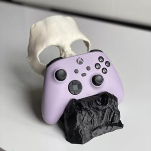 Può includere: Un controller Xbox lilla poggia su una base nera strutturata, con un teschio bianco sullo sfondo. Il controller presenta pulsanti neri e un D-pad. Tema di gioco.