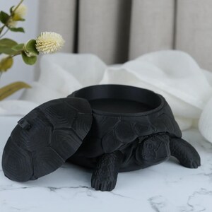 Könnte beinhalten: Ein mattschwarzer Behälter in Schildkrötenform mit abnehmbarem Panzerdeckel. Die Schildkröte hat eine texturierte, schuppige Oberfläche an Kopf, Beinen und Panzer. Der Deckel liegt daneben und gibt den Blick auf ein rundes, hohles Innenfach frei.
