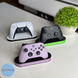 May include: Three gaming console controllers: white, black, and lavender. Each controller rests on a matching base with a contrasting color trim. The controllers are arranged on a white surface, with a small green plant in the background.