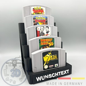 Könnte beinhalten: Ein schwarzer Plastikständer mit fünf Steckplätzen für Nintendo 64-Spielkassetten. Die Kassetten sind mit "Pokemon Snap", "Pokemon Stadium", "FIFA Soccer 64", "Donkey Kong 64" und "The Legend of Zelda: Ocarina of Time" beschriftet. Der Ständer trägt ein Etikett mit der Aufschrift "WUNSCHTEXT MADE IN GERMANY".