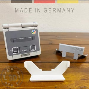 Könnte beinhalten: Eine grau-weiße Nintendo Super Nintendo Entertainment System (SNES) Konsole mit passendem grau-weißem Controller und zwei weißen Ständern. Der Text "MADE IN GERMANY" befindet sich über der Konsole.