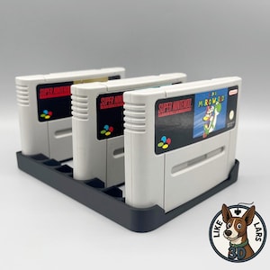 Könnte beinhalten: Ein schwarzer, 3D-gedruckter Patronenhalter mit drei Super Nintendo-Spielpatronen. Die Patronen sind hellgrau mit farbenfrohen Etiketten. Eine Patrone zeigt eine "Mario World"-Grafik. Der Halter hat einzelne Schlitze für jedes Spiel.