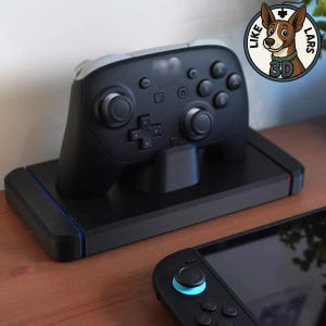 Könnte beinhalten: Schwarzer und grauer Videospiel-Controller auf einer schwarzen Ladestation. Der Controller hat mehrere Tasten und Joysticks. Eine zweite Spielkonsole ist im Vordergrund zu sehen. Das Bild enthält ein Logo mit einem Hund.