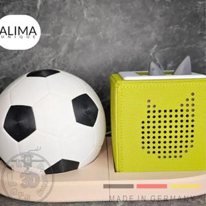 Pode incluir: Uma bola de futebol branca e preta ao lado de um alto-falante verde limão com um design de rosto de gato. O alto-falante tem dois detalhes cinza em forma de barbatana de tubarão. Os itens estão em uma base de madeira clara. O texto "MADE IN GERMANY" é visível.