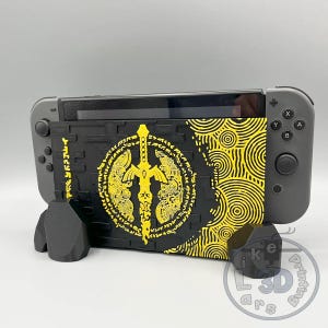 Op de afbeelding: Een zwart-gele decoratieve hoes voor een Nintendo Switch. De hoes heeft een geel zwaardontwerp op een zwarte achtergrond, met een bijpassende standaard. De Switch is grijs.
