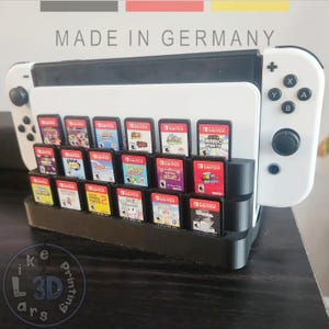 Puede incluir: Una consola Nintendo Switch blanca y negra con un soporte negro para cartuchos de juegos. El soporte está lleno de varios cartuchos de juegos. El texto "MADE IN GERMANY" está en la parte superior.
