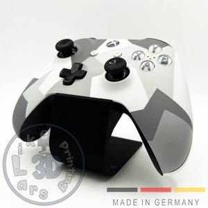 Könnte beinhalten: Ein kabelloser Videospiel-Controller mit einem geometrischen Tarnmuster in Weiß, Hellgrau und Dunkelgrau. Der Controller verfügt über zwei schwarze Analogsticks, ein schwarzes Steuerkreuz und silberne Aktionstasten mit farbigen Markierungen.