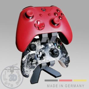 Op de afbeelding: Een rode Xbox-controller staat bovenop een zwart-witte controller, beide ondersteund door een zwarte standaard. De controllers staan tegen een grijze achtergrond. De tekst "MADE IN GERMANY" is zichtbaar.