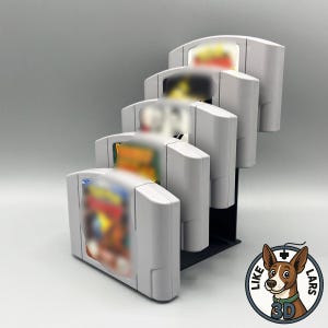 Könnte beinhalten: Ein schwarzer, gestufter Kunststoff-Displayständer, der fünf hellgraue Videospielkassetten in einer vertikalen, treppenartigen Anordnung hält. Die Kassetten haben unscharfe Etiketten auf der Vorderseite und der Ständer hat eine stabile Basis.