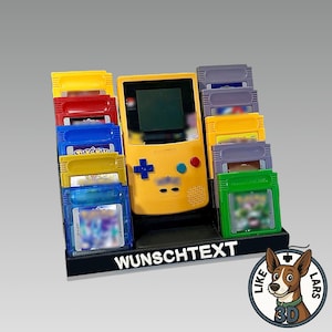 Könnte beinhalten: Eine gelbe Game Boy-Konsole und farbenfrohe Spielmodule werden auf einem schwarzen Ständer präsentiert. Die Module sind in verschiedenen Farben gehalten, darunter Rot, Blau und Gelb. Der Ständer trägt den Text "WUNSCHTEXT".