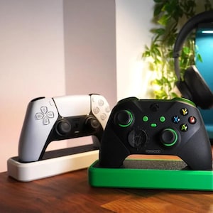 Op de afbeelding: Twee gamecontrollers, één wit en één zwart, op standaards. De witte controller heeft een symmetrische knoppenindeling. De zwarte controller heeft groene accenten en het woord "FOXWOOD" is erop gedrukt. De controllers staan op een houten oppervlak.