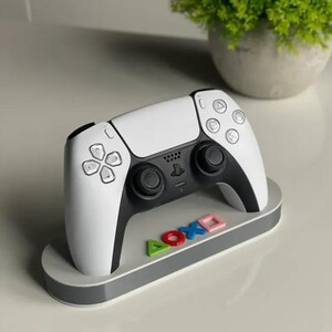 Könnte beinhalten: Ein weiß-schwarzer PlayStation 5-Controller auf einem grauen Ständer mit farbenfrohen, knopfförmigen Akzenten. Der Ständer hat die Buchstaben A, O, X und ein Quadrat. Der Controller hat eine schwarze Mitte und weiße Seiten.