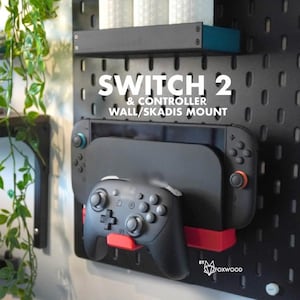Peut inclure: Support mural noir pour Switch 2 et manette. Le support est fixé à une plaque perforée noire. La Switch 2 est noire avec des touches de rouge et de bleu. La manette est noire et repose sur un support rouge.