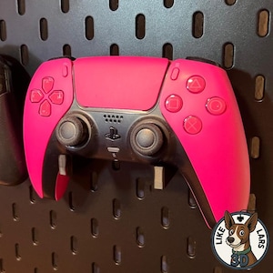 Könnte beinhalten: Ein leuchtend pinkfarbener kabelloser Videospiel-Controller mit schwarzen Analogsticks, einem schwarzen Mittelteil mit PlayStation-Logo und Standard-Aktionstasten wie Dreieck, Kreis, Kreuz und Quadrat, montiert an einer schwarzen Lochwand.