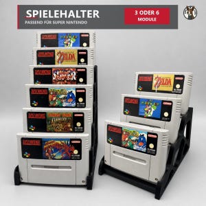 Könnte beinhalten: Zwei schwarze Kunststoff-Spielhalterungen für Super Nintendo-Module. Der linke Ständer hält sechs Module, der rechte drei. Der Text oben lautet SPIELEHALTER PASSEND FÜR SUPER NINTENDO und 3 ODER 6 MODULE.