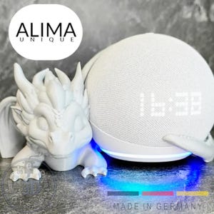 Pode incluir: Um alto-falante inteligente branco com um visor de relógio digital, apoiado em um suporte branco em forma de dragão. O suporte tem uma luz LED azul na base. O texto "ALIMA UNIQUE" está em um círculo acima. "MADE IN GERMANY" na parte inferior.