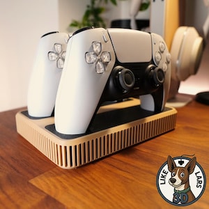 Op de afbeelding: Een wit en zwarte gamecontroller en twee witte gamecontrollers rusten op een beige oplaadstation. Het oplaadstation heeft een geribbeld ontwerp. De controllers hebben zwarte joysticks en doorzichtige knoppen.