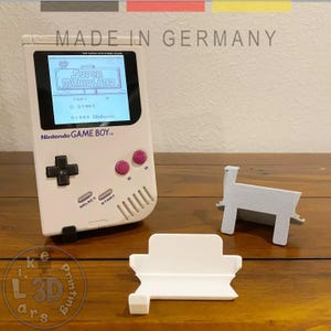 Peut inclure: Une console portable Nintendo Game Boy blanche avec un écran affichant "Super Marioland". La console a un pavé directionnel noir, des boutons roses et le texte "MADE IN GERMANY". Deux supports blancs et gris sont à côté de la console.