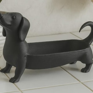 Könnte beinhalten: Eine mattschwarze Schale aus Gussmetall in Form eines Dackels. Die Schale verfügt über einen langen, hohlen Körper zur Aufbewahrung kleiner Gegenstände, wobei Kopf und Schwanz des Hundes an beiden Enden nach oben ragen. Die Oberfläche ist texturiert.