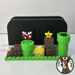 May include: A black Nintendo Switch dock with 3D-printed Super Mario Bros. themed accessories. Includes a Piranha Plant, a yellow question mark block, a star, and green pipes. The accessories sit on a green grass base.