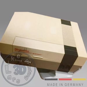 Könnte beinhalten: Eine beige Nintendo Entertainment System Konsole mit schwarzen Akzenten. Das Nintendo-Logo und die Worte "Entertainment System" sind rot. Die Konsole steht auf einem transparenten Ständer. Der Text "MADE IN GERMANY" ist unten zu sehen.