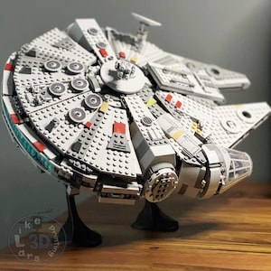 Puede incluir: Un modelo detallado de la nave espacial Millennium Falcon, construida con ladrillos LEGO. El modelo es gris y blanco con detalles rojos, amarillos y negros. La nave espacial está sobre un soporte negro.