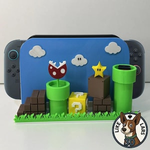 Könnte beinhalten: Ein blauer Nintendo Switch-Ständer mit einer 3D-gedruckten Super Mario Bros.-Szene. Die Szene umfasst eine rot-weiße Piranha-Pflanze, einen gelben Fragezeichenblock, einen braunen Sternblock, grüne Rohre und weiße Wolken.