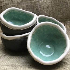 Puede incluir: Cuatro cuencos de cerámica con un exterior gris oscuro y un interior verde claro. Los cuencos tienen un borde blanco y una apariencia ligeramente irregular, hecha a mano.