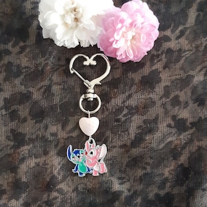 以下が含まれることがあります： ハート型のチャームにアニメのカップルが描かれたシルバーのキーホルダー。チャームはシルバーのスプリングクラスプに付いています。キーホルダーはピンクと白の花に囲まれています。