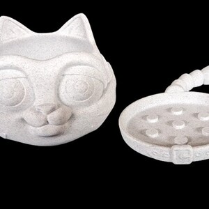 May include: A white ceramic cat-shaped dish with a matching tail and a round base with small indentations. The cat's face has large eyes and a smiling mouth.