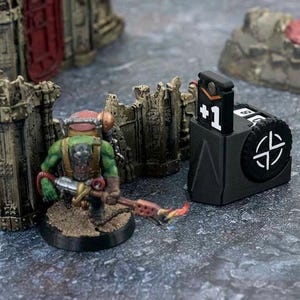 May include: Miniature figure of a green-skinned orc with a weapon, standing near a black game die with a white crosshair symbol. The scene includes miniature terrain pieces, suggesting a tabletop war game setting.