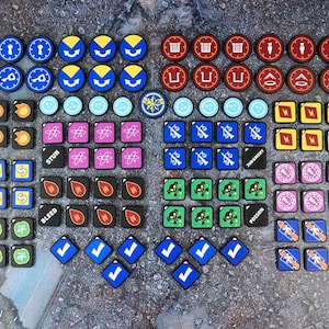 Puede incluir: Una colección de fichas de juego variadas en formas circulares, cuadradas y de diamante. Las fichas presentan diversos colores, incluyendo azul, rojo, amarillo, morado, verde y negro, con iconos impresos que representan símbolos y efectos.