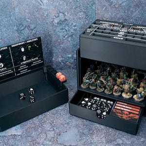 May include: A black plastic tabletop gaming organizer with a hinged lid featuring printed game stats, equipment, and strategy labels. The unit includes a dice tray with black and red dice, a storage compartment for miniature figures, and a pull-out drawer.