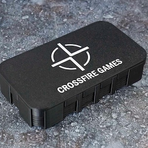 Könnte beinhalten: Ein rechteckiger schwarzer Aufbewahrungskoffer mit abgerundeten Ecken, der auf dem Deckel den weißen Schriftzug CROSSFIRE GAMES und ein weißes kreisförmiges Fadenkreuz-Logo trägt. Der Koffer hat eine matte, texturierte Oberfläche und gerippte Seiten.