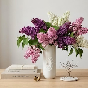 May include: A white ceramic vase filled with a vibrant bouquet of lilacs in shades of purple, pink, and white. The vase sits on a wooden surface with books and a decorative tree sculpture.