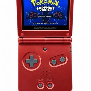 May include: A metallic red Game Boy Advance SP handheld gaming console with a flip-top screen. The screen displays the Pokémon Sapphire Version title screen with the text Press Start and 2003 Game Freak inc. visible below the logo.
